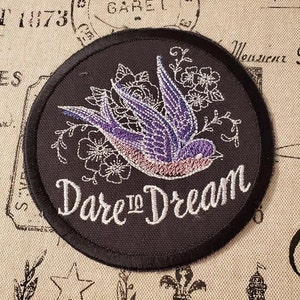 May include: A black embroidered patch with a purple and pink bird flying with flowers and the text "Dare to Dream" in white.