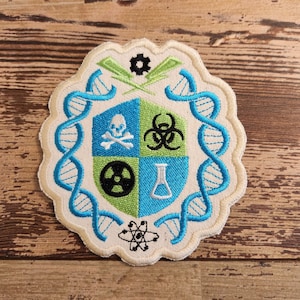 May include: A white embroidered patch with a blue and green shield featuring symbols of science, including a skull and crossbones, a biohazard symbol, a radiation symbol, a beaker, and a DNA helix.