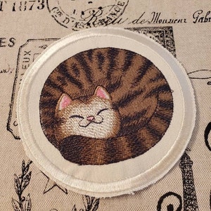 May include: A white embroidered patch with a brown tabby cat curled up sleeping. The cat has a white face and pink ears.
