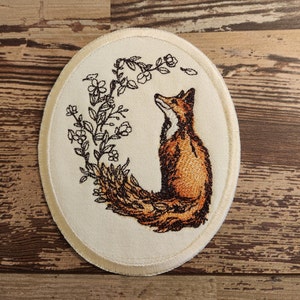 May include: A white oval embroidered patch with a brown fox sitting with its tail curled around a floral design. The fox is looking up and the flowers are in bloom.