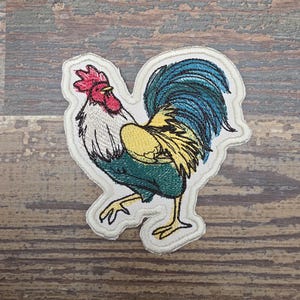May include: Embroidered patch featuring a colorful rooster. The rooster has a red comb and wattle, white neck, yellow and green body, and a blue tail. The patch is outlined with a cream-colored border.