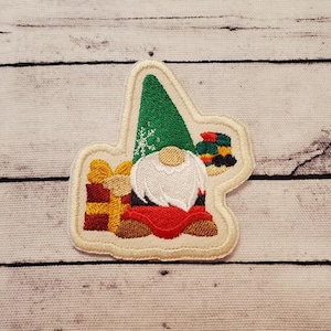 May include: Embroidered patch featuring a Christmas gnome with a white beard, red suit, and green hat. The gnome is surrounded by wrapped gifts in gold, red, and green. The patch has a cream-colored border and is set against a light wood-grain background.