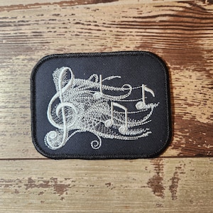 May include: A black embroidered patch with a white musical note design. The design features a treble clef and several musical notes.