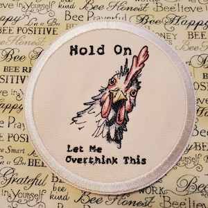 May include: White round patch with a white border. The patch features a cartoon chicken with a red comb and wattle. The text "Hold On" is above the chicken and the text "Let Me Overthink This" is below the chicken.
