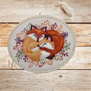 May include: Embroidered patch featuring two hugging foxes in shades of orange, brown, and white, surrounded by autumn leaves. The patch is circular with a beige border, perfect for adding to clothing or accessories.