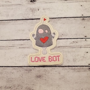 May include: Embroidered iron-on patch featuring a gray robot with a pink heart and the text "LOVE BOT" in pink.