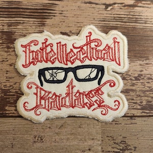 May include: A white embroidered patch with red stitching. The patch features the words "Intellectual Badass" in a stylized font. A pair of black, cracked glasses are centered above the text.