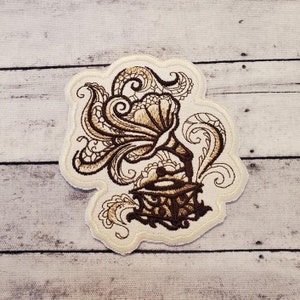 May include: A brown embroidered patch featuring a vintage-style record player with intricate floral designs. The record player is surrounded by swirling patterns and has a detailed design.