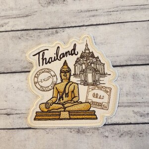 May include: Embroidered patch with a gold and brown design featuring a seated Buddha, a temple, and the word "Thailand". The patch also includes a stamp with the words "Bangkok" and "Thailand".