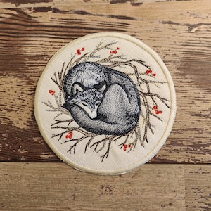 May include: Embroidered patch featuring a gray fox curled up asleep in a nest of branches with red berries. The patch is circular and has a white background.
