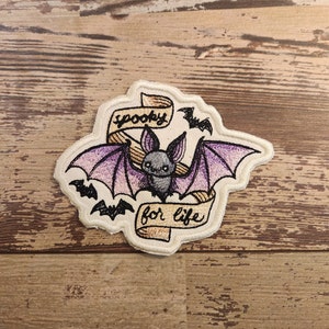 May include: Embroidered patch with a purple bat with black eyes and wings, a banner that reads "spooky for life" and two smaller bats.