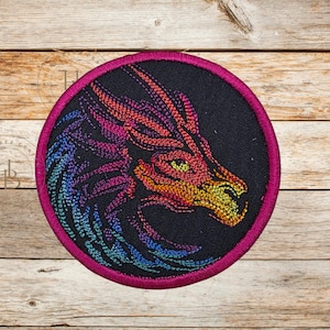 May include: Embroidered patch featuring a dragon's head in profile. The dragon is rendered in vibrant colors, including red, orange, yellow, blue, and purple, against a black background. The patch has a pink border.