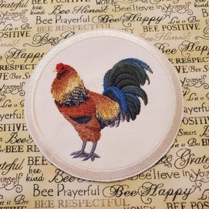 May include: A white round patch with a colorful embroidered rooster design. The rooster is facing right with a red comb and wattle, brown and yellow feathers, and a blue and green tail.