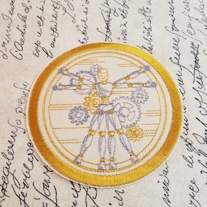 May include: A circular embroidered patch with a gold border. The center of the patch features a white and gray image of a human figure with gears and cogs for limbs. The image is reminiscent of Leonardo da Vinci's Vitruvian Man.