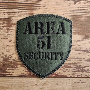 May include: A green fabric patch with black embroidered text that reads "AREA 51 SECURITY".