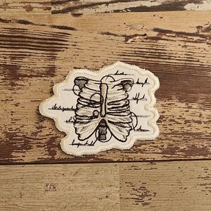 May include: A beige embroidered patch with a detailed anatomical illustration of a rib cage. The patch has a white background with black lines and text.