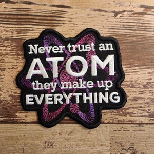 May include: A black embroidered patch with a purple and white atom design. The text reads "Never trust an ATOM they make up EVERYTHING".