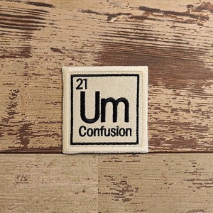 May include: A beige fabric patch with black embroidery. The patch is shaped like a square and has a black border. The text "Um" is in the center of the patch, with the number "21" above it. The text "Confusion" is below the word "Um".