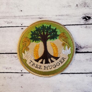 May include: A round, green and brown embroidered patch with a tree, wheat stalks, and a banner that reads "Tree Hugger".