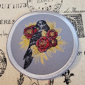 May include: A gray embroidered patch with a black crow surrounded by red roses and gold leaves.