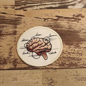 May include: A round, off-white embroidered patch featuring a detailed illustration of a brain in shades of brown and tan. The brain is labeled with black lines and text. The patch has a cream-colored border.