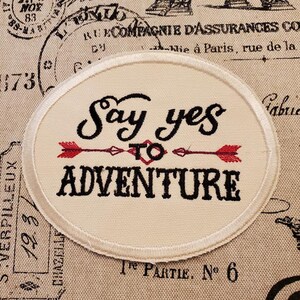 May include: White fabric patch with a black embroidered saying "Say yes to adventure" with two red arrows pointing to the right.
