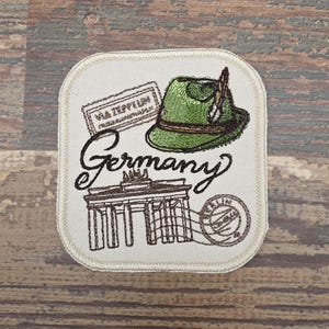 May include: Embroidered patch featuring a green hat with a feather, the word "Germany" in script, and a depiction of the Brandenburg Gate. Includes a stamp that says "Via Zeppelin". The patch is on a cream-colored background.