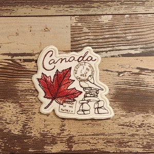 May include: Embroidered patch with a red maple leaf, a vintage-style stamp that says "Canada" and a drawing of a stack of stones. The patch is beige with brown stitching.