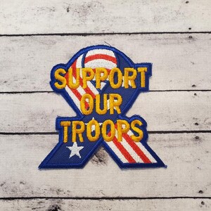 May include: A blue and red ribbon with white stars and stripes, with the words "Support Our Troops" in yellow lettering.