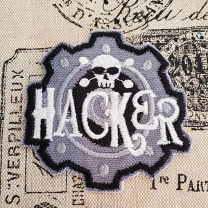 May include: A gray and black embroidered patch with a skull and crossbones design and the word "HACKER" in white lettering.