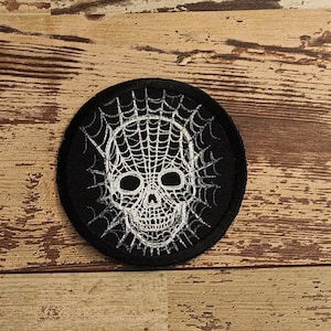 May include: A black and white embroidered patch with a skull design. The skull is surrounded by a spider web.
