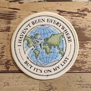 Puede incluir: Parche bordado redondo con el texto "I HAVEN'T BEEN EVERYWHERE BUT IT'S ON MY LIST". El parche presenta un mapa del mundo azul y verde con un avión gris. El parche tiene un borde color crema.