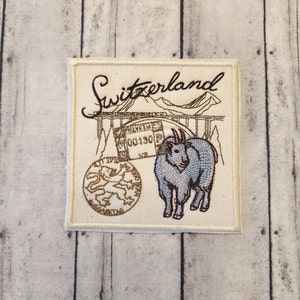 May include: A white embroidered patch with the word "Switzerland" in cursive script. The patch features a mountain scene with a bridge, a goat, and a circular emblem with a lion.