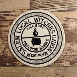 May include: Circular patch with the text "Salem Local Witches Union" in black on an off-white background. The patch features a cauldron, flame, and the text "Fire Burn Cauldron Bubble" and "Est. 1692". A black Greek key border surrounds the design.