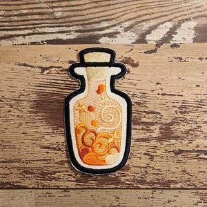 May include: A yellow and orange embroidered patch of a glass bottle filled with a swirling liquid. The bottle has a cork stopper and a black outline.