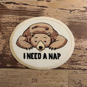 May include: A beige embroidered patch with a brown bear sleeping on its belly. The text "I NEED A NAP" is below the bear.