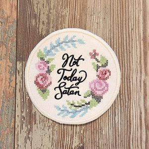 May include: A round, off-white patch with the phrase "Not Today Satan" in black script. The text is surrounded by a cross-stitched floral wreath in pink, blue, and green. The patch has a cream-colored border.