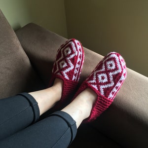 May include: A pair of red knitted slippers with a white geometric pattern. The slippers are worn by a person sitting on a brown couch.