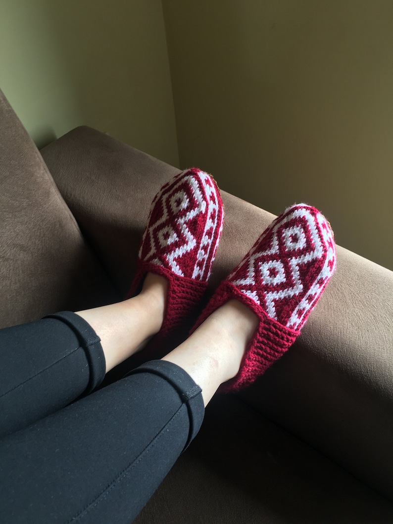 May include: A pair of red knitted slippers with a white geometric pattern. The slippers are worn by a person sitting on a brown couch.