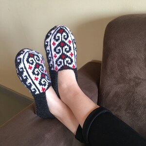 May include: A pair of black and white knitted slippers with red accents. The slippers have a geometric pattern and are worn by a person sitting on a brown couch.