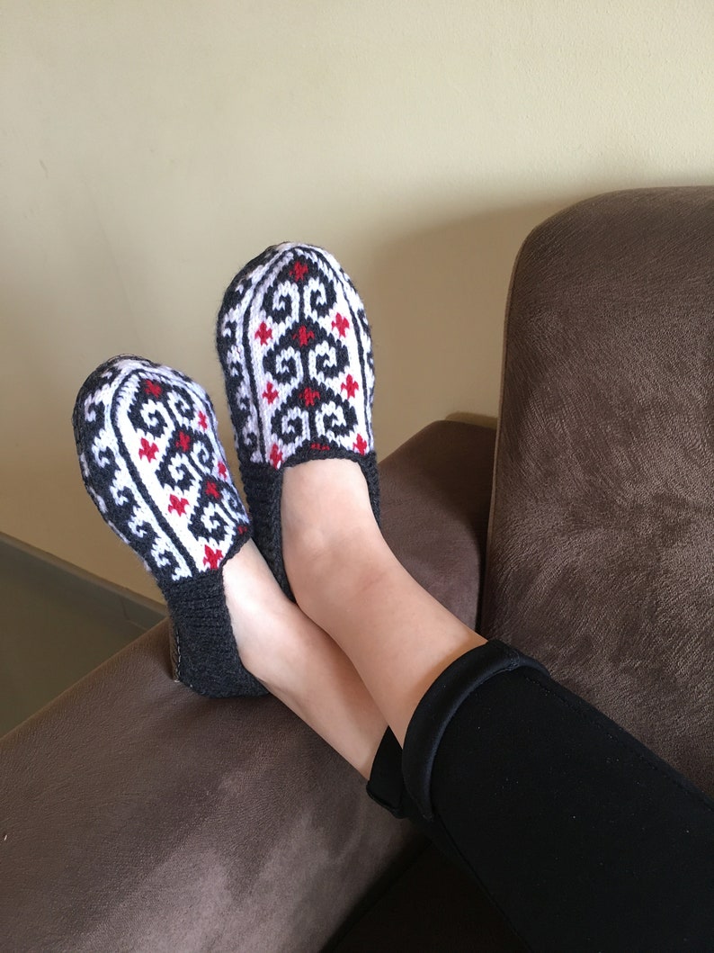 May include: A pair of black and white knitted slippers with red accents. The slippers have a geometric pattern and are worn by a person sitting on a brown couch.