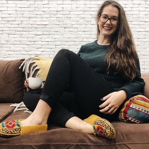 May include: A woman wearing black pants and a green sweater sits on a brown couch with a patterned throw pillow. She is holding a white mug and wearing yellow knitted slippers with colorful patterns.