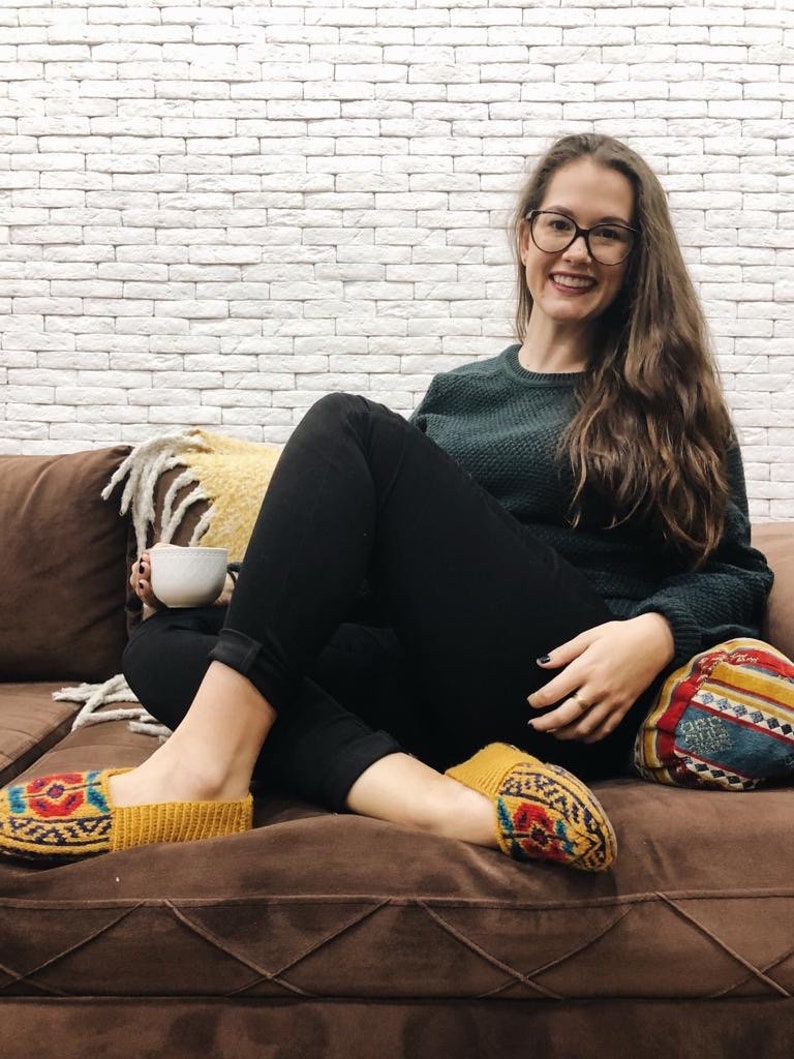 May include: A woman wearing black pants and a green sweater sits on a brown couch with a patterned throw pillow. She is holding a white mug and wearing yellow knitted slippers with colorful patterns.