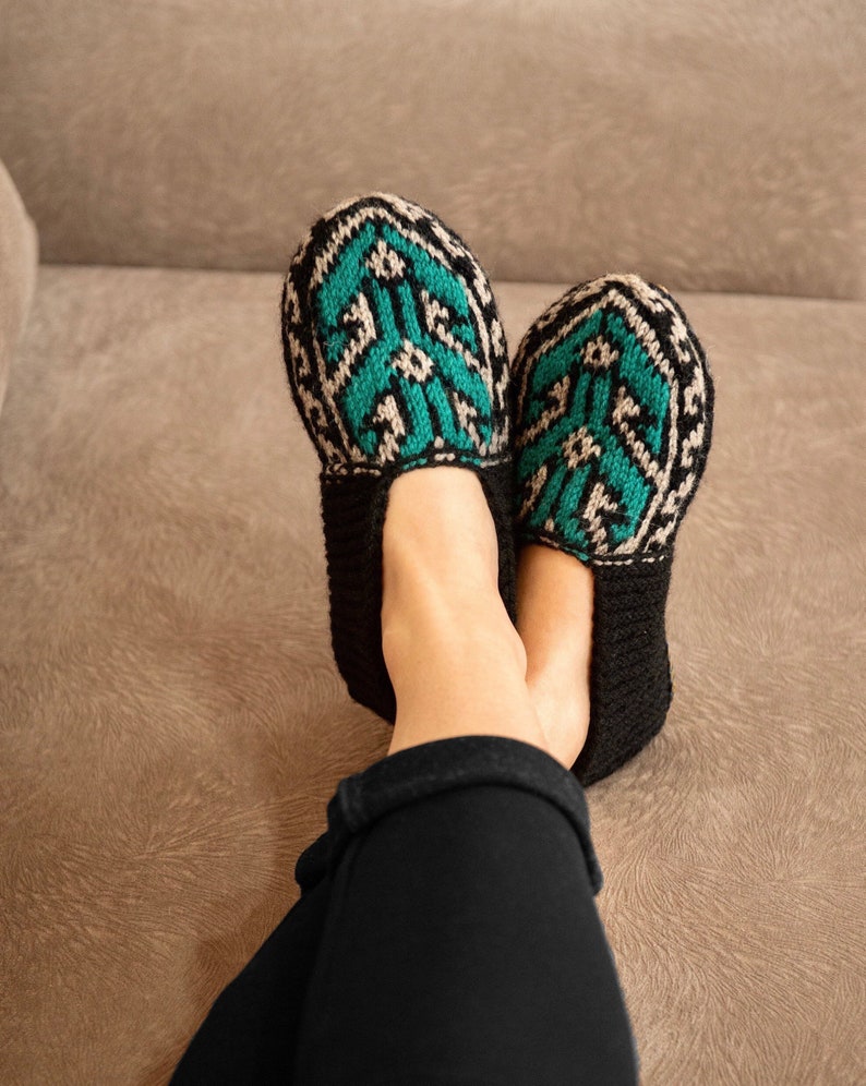 May include: A pair of black knitted slippers with a green and white geometric pattern. The slippers are worn by a person sitting on a brown couch.