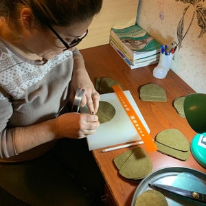 May include: A person is working on a craft project, cutting out pieces of brown leather with a small tool. There are several stacks of leather pieces on a table, along with a ruler, a pencil, and a pair of scissors.