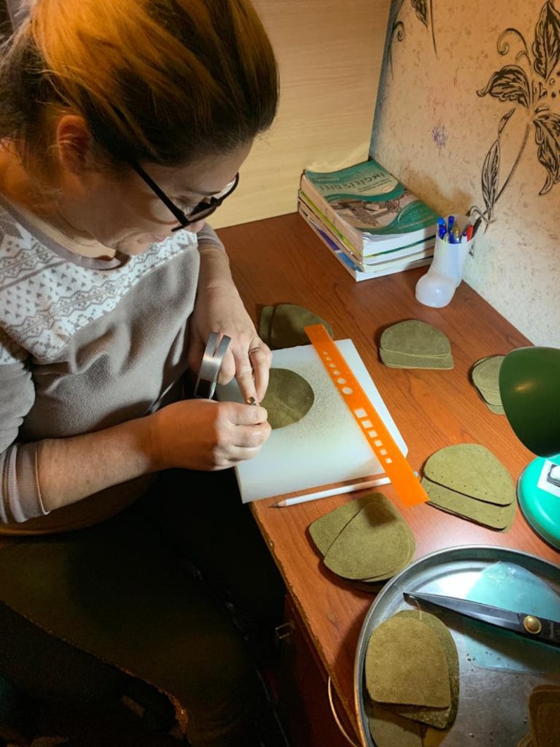 May include: A person is working on a craft project, cutting out pieces of brown leather with a small tool. There are several stacks of leather pieces on a table, along with a ruler, a pencil, and a pair of scissors.