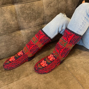 May include: A pair of red, green, and black patterned wool socks with intricate designs. The socks are worn on a person's feet and are resting on a brown couch.