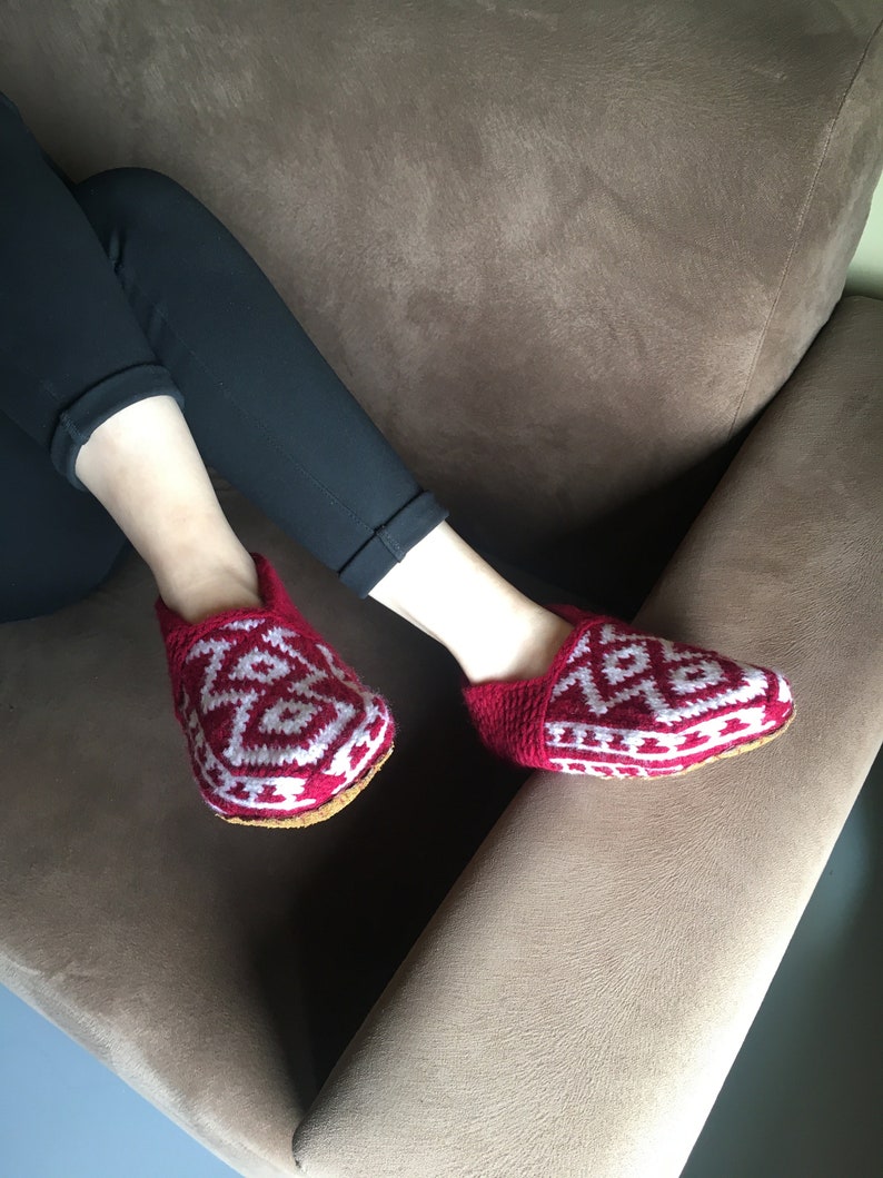 May include: Red and white knitted slippers with a geometric pattern. The slippers are worn by a person sitting on a brown couch.