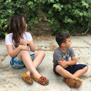 May include: Two people are sitting on a stone patio. The woman is wearing a white t-shirt, blue and white patterned shorts, and yellow and red embroidered slippers. The boy is wearing a gray t-shirt, blue shorts, and yellow and red embroidered slippers.