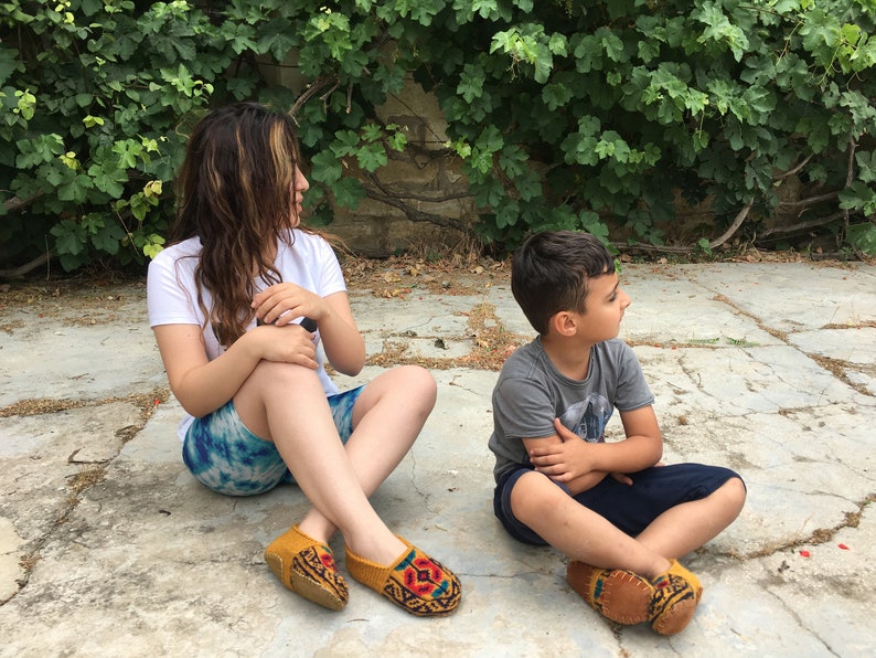 May include: Two people are sitting on a stone patio. The woman is wearing a white t-shirt, blue and white patterned shorts, and yellow and red embroidered slippers. The boy is wearing a gray t-shirt, blue shorts, and yellow and red embroidered slippers.